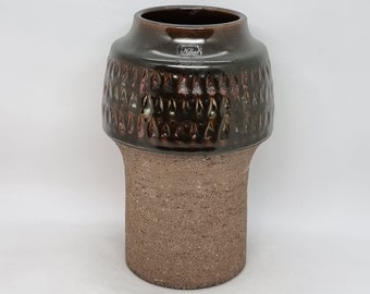 May include: A brown ceramic vase with a textured, geometric pattern. The vase has a wide, round base and a narrower neck. The vase is made of a rough, textured material.