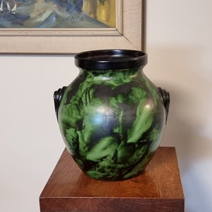 May include: A green and black ceramic vase with two handles. The vase has a mottled green glaze with black accents. It is sitting on a wooden pedestal.