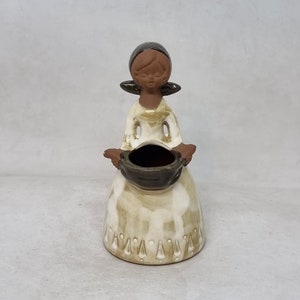 May include: A ceramic figurine of a woman in a white dress and brown headscarf holding a brown pot. The figurine has a rustic, hand-painted look.