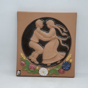 Puede incluir: Un azulejo de terracota con un diseño en relieve de una pareja bailando. La pareja está rodeada por un círculo de esmalte marrón y un borde de flores y espigas de trigo. El azulejo está firmado "JIC" en la esquina inferior izquierda.