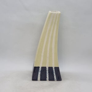 May include: A tall, cream-coloured ceramic vase with a striped design. The vase has a narrow opening and a wide base. The base is decorated with a band of dark purple and white stripes.