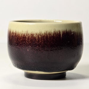 Puede incluir: Un pequeño cuenco redondo de cerámica con un esmalte marrón y crema. El cuenco tiene un borde ligeramente acampanado y una base redondeada.