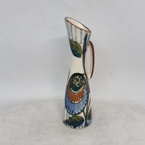 Peut inclure: Un petit vase en céramique blanche avec une anse brune. Le vase est décoré d'un motif floral bleu, vert et orange.