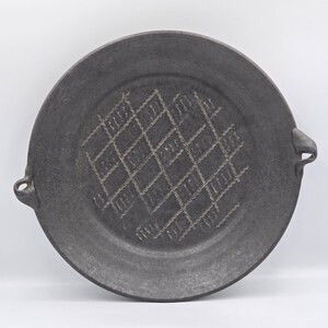 Puede incluir: Una bandeja redonda de cerámica gris oscuro con dos pequeñas asas laterales integradas. El centro de la bandeja presenta un patrón de rombos texturizado y en relieve, formado por líneas que se cruzan con un detalle decorativo ondulado claro.
