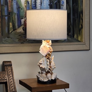 May include: A table lamp featuring a base constructed from a cluster of various white and tan seashells. The lamp is topped with a cylindrical, off-white fabric lampshade. A small, rectangular, light-colored paper tag hangs from the seashell base.