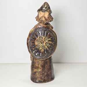 Puede incluir: Una figurita de cerámica de un guerrero con barba, que lleva un casco y sostiene un escudo con un diseño circular. La figurita es marrón y verde con un acabado texturizado.