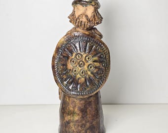 May include: A ceramic figurine of a warrior with a beard, wearing a helmet and holding a shield with a circular design. The figurine is brown and green with a textured finish.