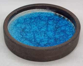 May include: A small, round ceramic dish with a dark brown rim and a bright blue interior. The blue glaze has a textured, crackled finish.