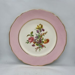 May include: A white ceramic plate with a pink rim and gold trim. The plate features a floral design with a pink rose, yellow iris, and other colourful flowers.