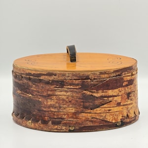 Può includere: Una scatola portaoggetti ovale realizzata in corteccia di betulla marrone testurizzata con un coperchio in legno liscio di colore chiaro. Il coperchio presenta una piccola maniglia verticale scura e semplici motivi a spirale disegnati a mano.