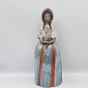 May include: A ceramic figurine of a woman in a long dress and bonnet, holding a bouquet. The dress is blue with red vertical stripes. The figurine has a vintage aesthetic.