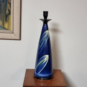 May include: A tall, blue ceramic candle holder with a black top and white and yellow abstract designs. The candle holder is on a wooden table.