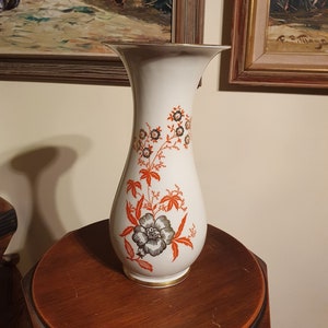 May include: A white ceramic vase with a floral design. The vase has a wide mouth and a narrow base. The floral design features orange, black, and gold accents.