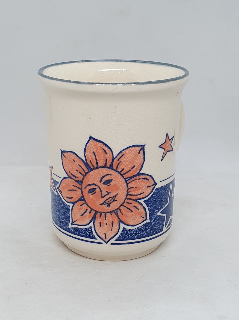 Puede incluir: Taza de cer&aacute;mica blanca con un dise&ntilde;o azul y naranja. El dise&ntilde;o presenta un sol con una cara y estrellas.