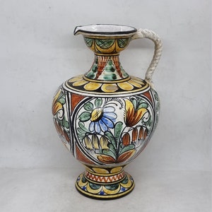 May include: A ceramic pitcher with a white base and a colorful floral design. The pitcher has a wide, round body and a narrow neck with a spout. The handle is white and attached to the neck of the pitcher.