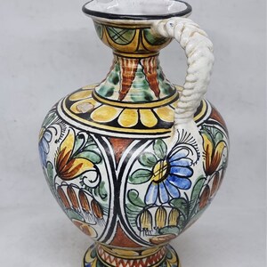 May include: A ceramic pitcher with a white, twisted handle and a colorful floral design. The pitcher has a round body with a wide base and a narrow neck. The design features yellow, blue, green, red, and brown colors.