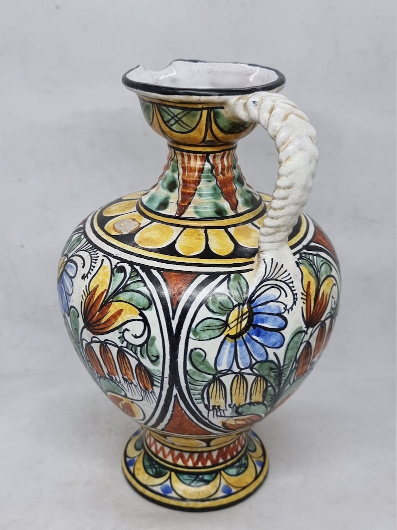 May include: A ceramic pitcher with a white, twisted handle and a colorful floral design. The pitcher has a round body with a wide base and a narrow neck. The design features yellow, blue, green, red, and brown colors.