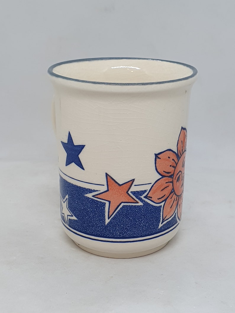 Puede incluir: Taza de cer&aacute;mica blanca con un dise&ntilde;o floral azul y naranja. El dise&ntilde;o presenta estrellas y flores.