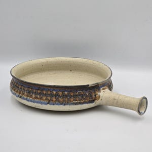 Op de afbeelding: Een ronde keramische koekenpan met één handvat, voorzien van een gespikkelde gebroken witte binnen- en buitenkant. De buitenrand is versierd met een band van donkerbruine en blauwe geometrische patronen, met een blauw accent op het uiteinde.
