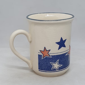 Puede incluir: Una taza de cer&aacute;mica blanca con un patr&oacute;n de estrellas azules y blancas. La taza tiene un borde azul y un asa.