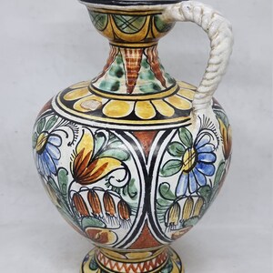 May include: A ceramic pitcher with a white, twisted handle. The pitcher is decorated with a colorful floral pattern in shades of blue, green, yellow, orange, and red.