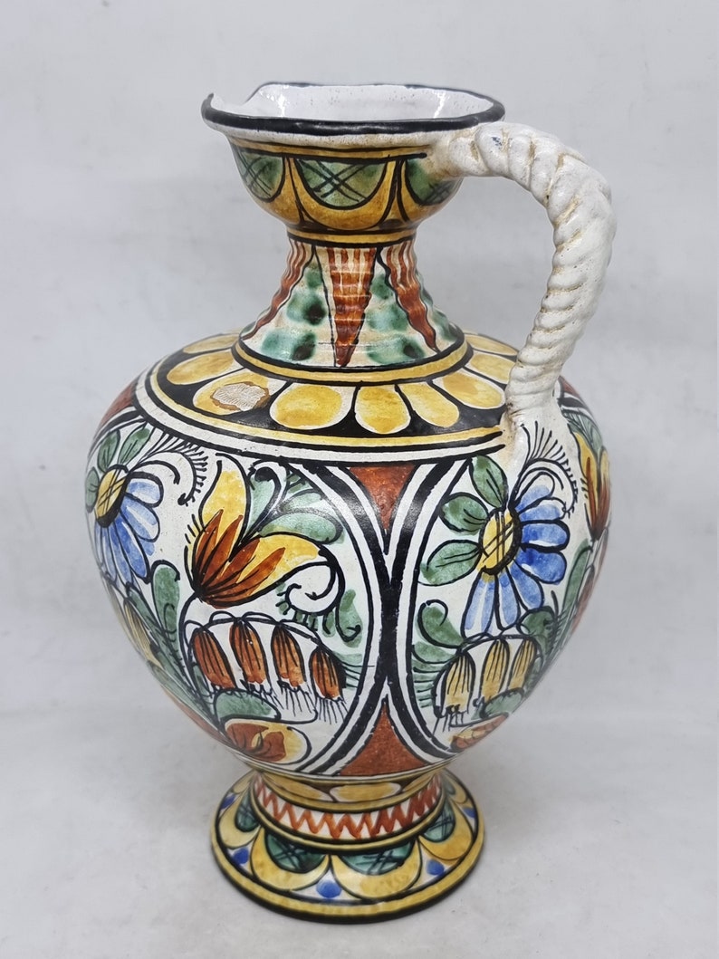 May include: A ceramic pitcher with a white, twisted handle. The pitcher is decorated with a colorful floral pattern in shades of blue, green, yellow, orange, and red.