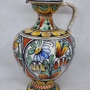 May include: A ceramic pitcher with a floral design in shades of yellow, orange, green, blue, and white. The pitcher has a wide base and a narrow neck with a handle.