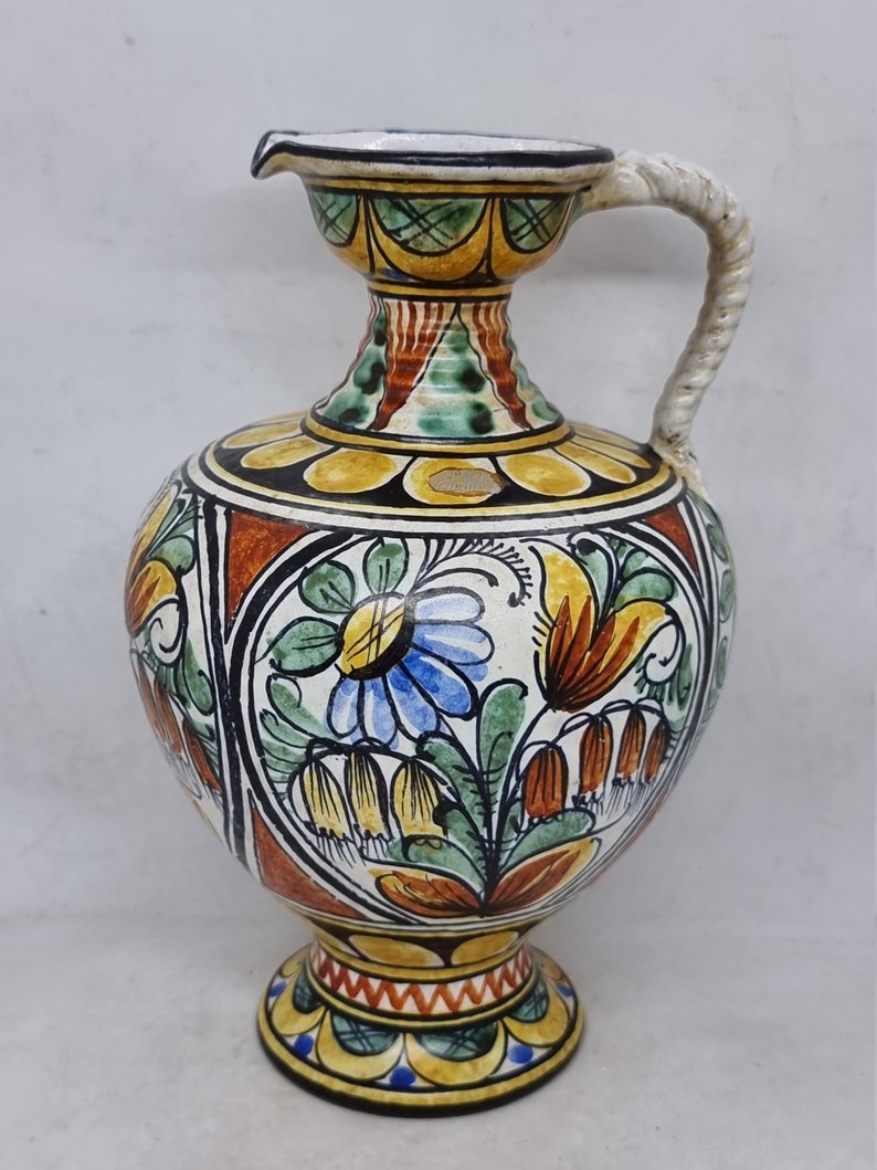 May include: A ceramic pitcher with a floral design in shades of yellow, orange, green, blue, and white. The pitcher has a wide base and a narrow neck with a handle.