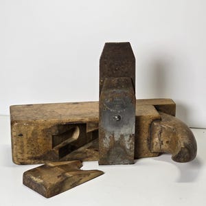 Peut inclure: Un rabot à main ancien en bois avec une lame en métal et une poignée incurvée. Le rabot est en bois et en métal patinés, montrant des signes d'âge et d'utilisation. Une petite pièce en bois est devant le rabot.