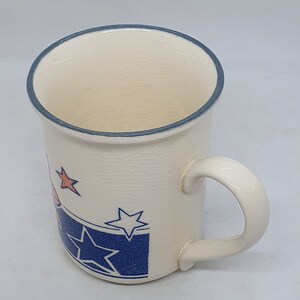 Puede incluir: Taza de cer&aacute;mica blanca con un dise&ntilde;o de estrellas azules y blancas y un borde azul. La taza tiene un asa y est&aacute; decorada con estrellas naranjas.