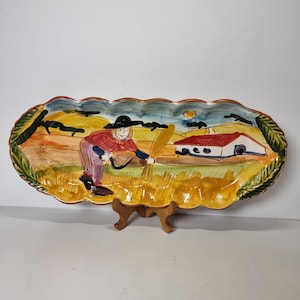 May include: A ceramic serving dish featuring a hand-painted scene of a person in a field with a house in the background. The dish has a scalloped edge and is decorated with green leaves.