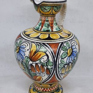 May include: A ceramic pitcher with a white, yellow, orange, green, and blue floral design. The pitcher has a twisted rope handle and a round base.