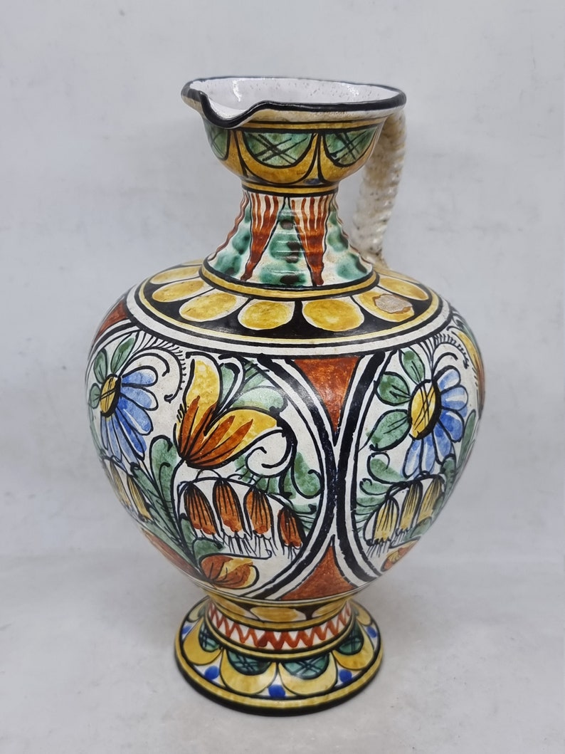 May include: A ceramic pitcher with a white, yellow, orange, green, and blue floral design. The pitcher has a twisted rope handle and a round base.