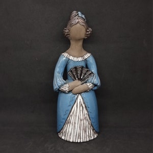May include: A ceramic figurine of a woman in a blue dress holding a black fan. The figurine has a white and brown striped skirt and a blue ribbon in her hair.