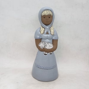 May include: A ceramic figurine of a girl wearing a light blue dress and a matching bonnet. She has blonde braids and brown skin. She is holding a white lamb in her arms.