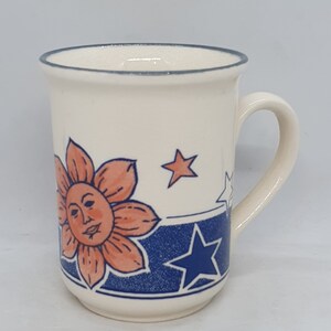Puede incluir: Una taza de cer&aacute;mica blanca con un dise&ntilde;o azul y naranja que presenta un sol con cara y estrellas. La taza tiene un asa.