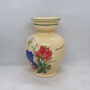 Può includere: Un vaso in ceramica color crema con un bordo verde e disegni floreali. Il vaso presenta un motivo floreale con fiori blu, rosa e bianchi. Il testo "Kleinsalzerhof" è stampato sul vaso.