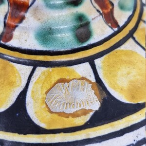 May include: Close-up of a ceramic pot with a yellow, black, and white geometric pattern. The pot has a silver sticker with the text "W.H. Handmade" on the bottom.