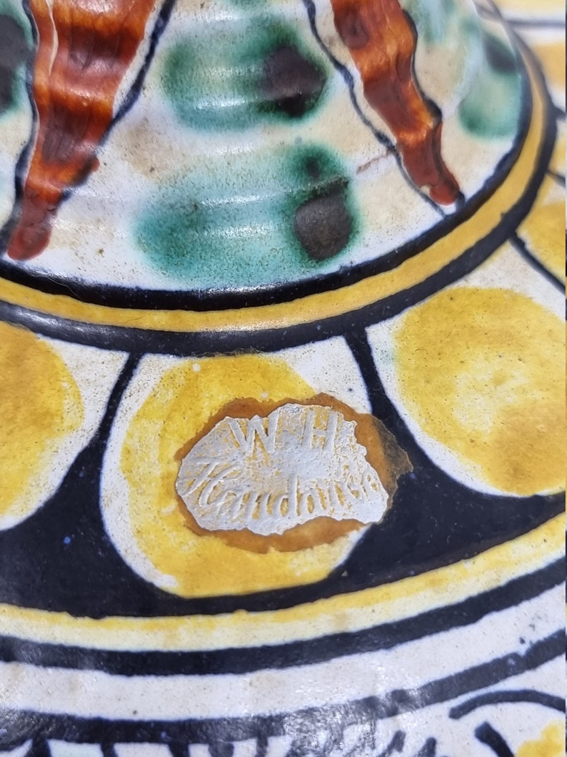 May include: Close-up of a ceramic pot with a yellow, black, and white geometric pattern. The pot has a silver sticker with the text "W.H. Handmade" on the bottom.