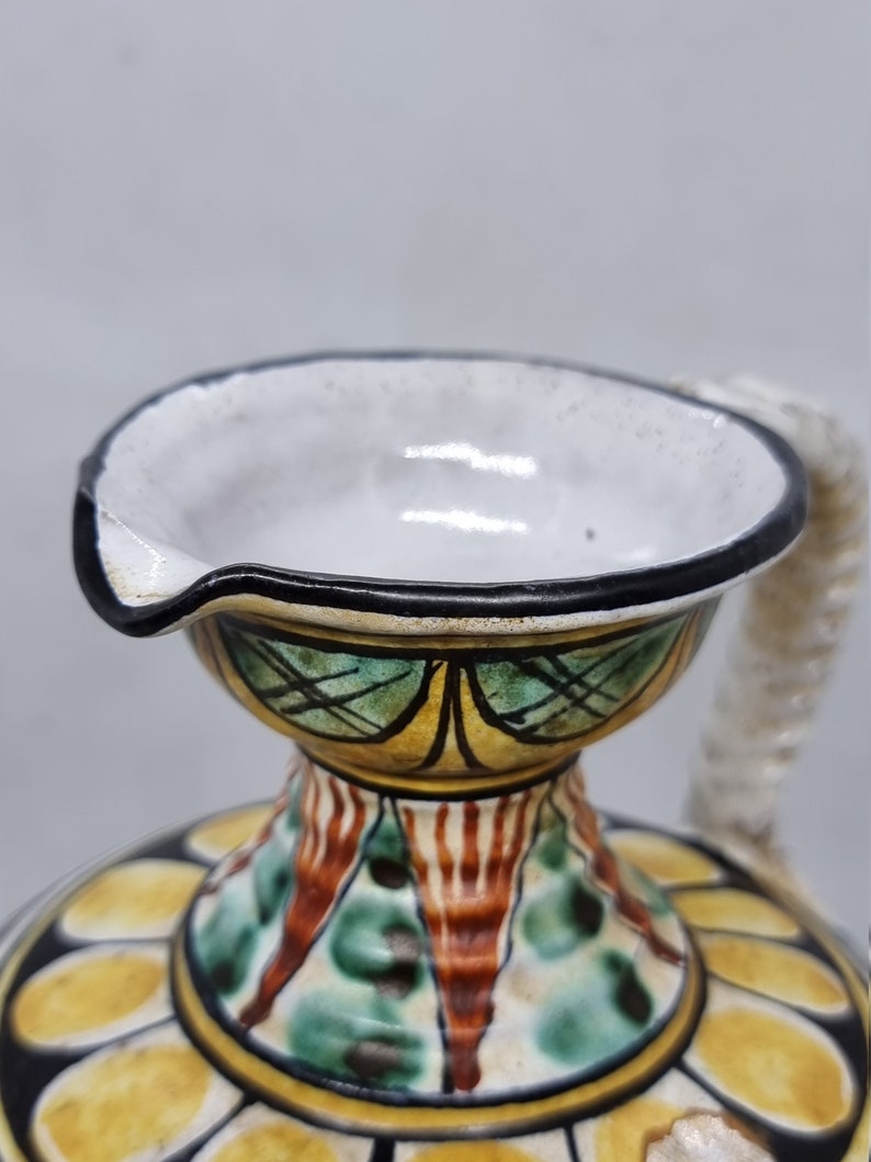 May include: A ceramic pitcher with a white glaze and a black rim. The pitcher has a floral design with yellow, green, and red accents. The pitcher has a spout and a handle.
