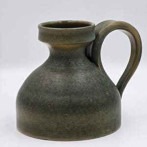 Peut inclure: Pichet en céramique vert foncé, fait à la main, avec un corps arrondi et une anse robuste. Le pichet a une large ouverture et un bord légèrement évasé. La surface présente une texture tachetée, lui donnant un aspect rustique. Idéal pour servir des boissons.