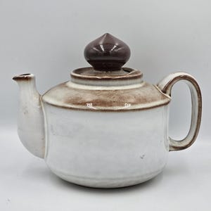 May include: A ceramic teapot featuring a white base with a brown glazed rim and a matching brown lid with a pointed knob. The teapot has a curved handle on the right side and a short spout on the left, with a smooth, glossy finish throughout.