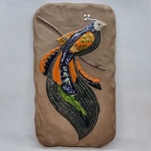 Puede incluir: Una placa de pared de cerámica con un pájaro estilizado con detalles en naranja, verde, azul y negro. El pájaro está posado en una rama sobre un fondo marrón.