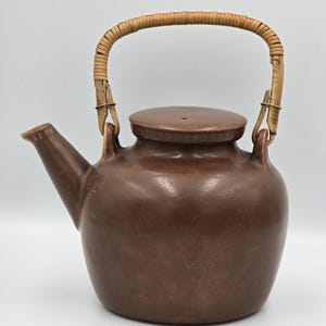 May include: A brown ceramic teapot with a smooth, matte finish, featuring a rounded body, a short spout, and a flat lid. The teapot has a curved, light-colored woven rattan handle attached to two small ceramic loops on the top of the vessel.