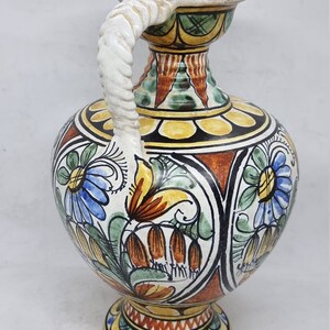 May include: A hand-painted ceramic vase with a twisted white handle. The vase is decorated with colorful floral patterns in shades of blue, green, yellow, orange, and red.