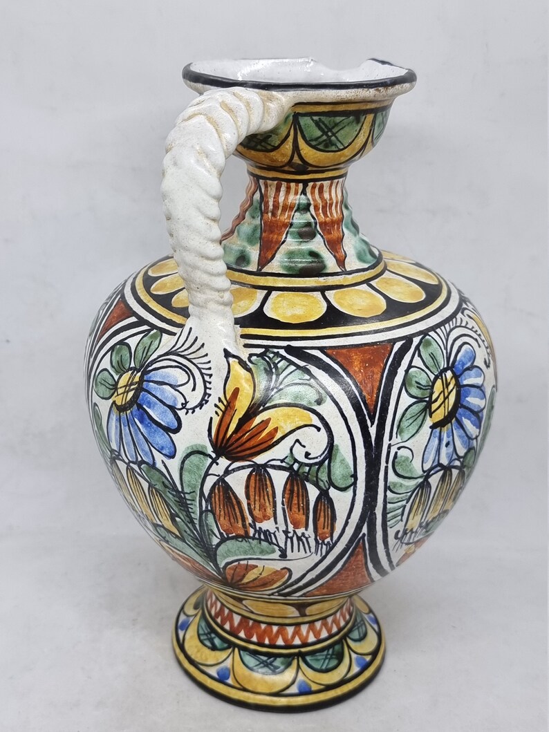 May include: A hand-painted ceramic vase with a twisted white handle. The vase is decorated with colorful floral patterns in shades of blue, green, yellow, orange, and red.