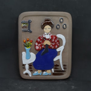 Puede incluir: Una placa de pared de cerámica que representa a una mujer con un vestido de lunares rojos y blancos sentada en una silla blanca, tejiendo. Tiene el pelo castaño y lleva zapatos azules. Hay un jarrón con flores naranjas sobre una mesa blanca a su lado.