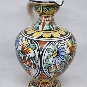May include: A hand-painted ceramic pitcher with a white background and colorful floral designs. The pitcher has a wide base and a narrow neck with a handle.