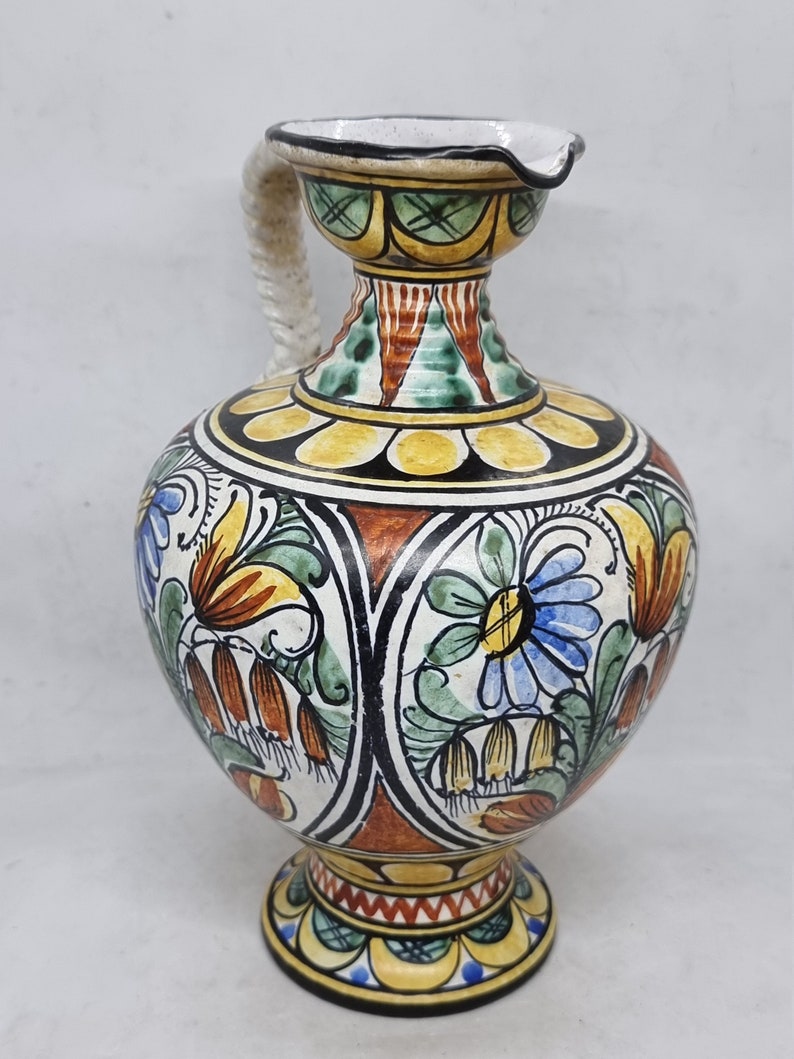 May include: A hand-painted ceramic pitcher with a white background and colorful floral designs. The pitcher has a wide base and a narrow neck with a handle.