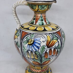 May include: A hand-painted ceramic pitcher with a white rope handle. The pitcher has a yellow, green, blue, and orange floral design.